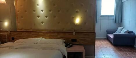 A hotel room with a large bed, a sofa, a bedside table, and a wall-mounted light fixture.