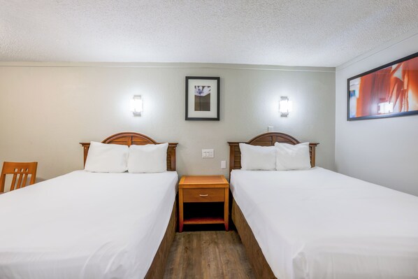 Habitación de hotel con dos camas individuales, una mesita de noche de madera entre ellas, una pequeña silla de madera y un cuadro enmarcado en la pared.