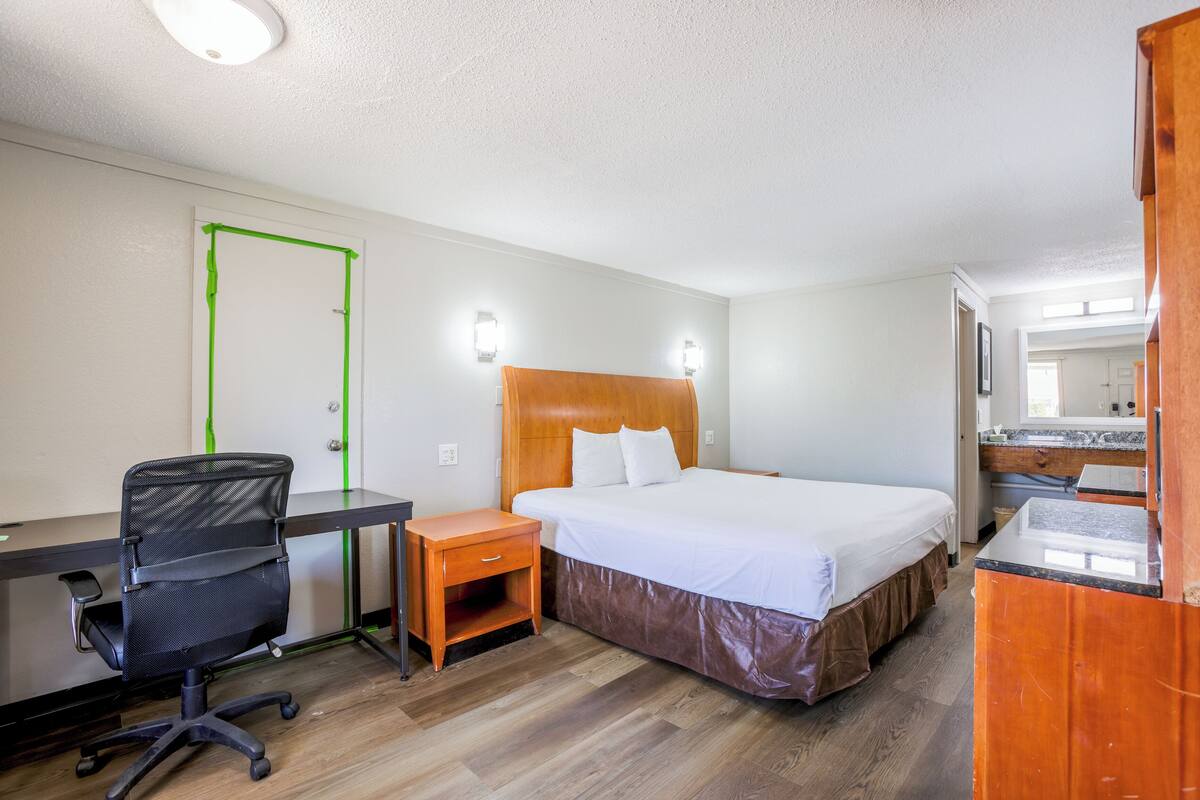A hotel room with a bed, desk, chair, and a small wooden nightstand.