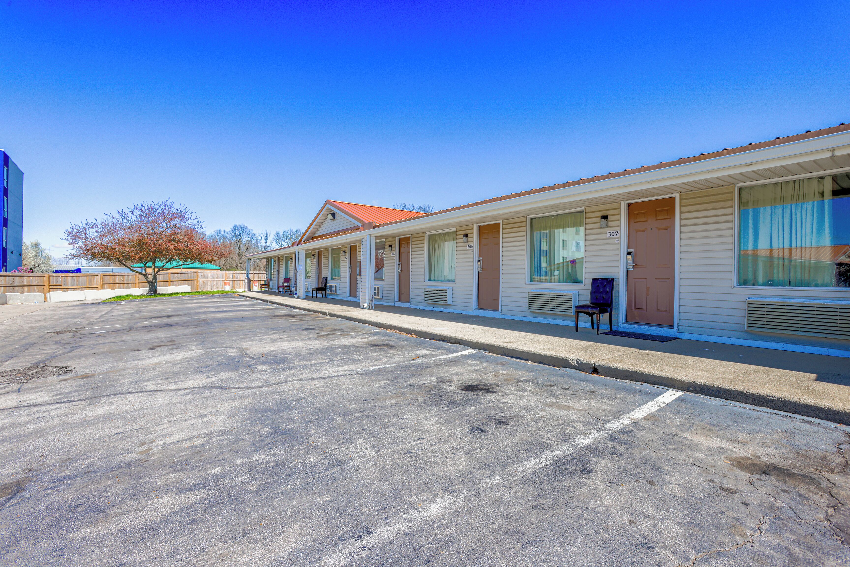A motel with a row of rooms, a parking lot, and a tree in the background.
