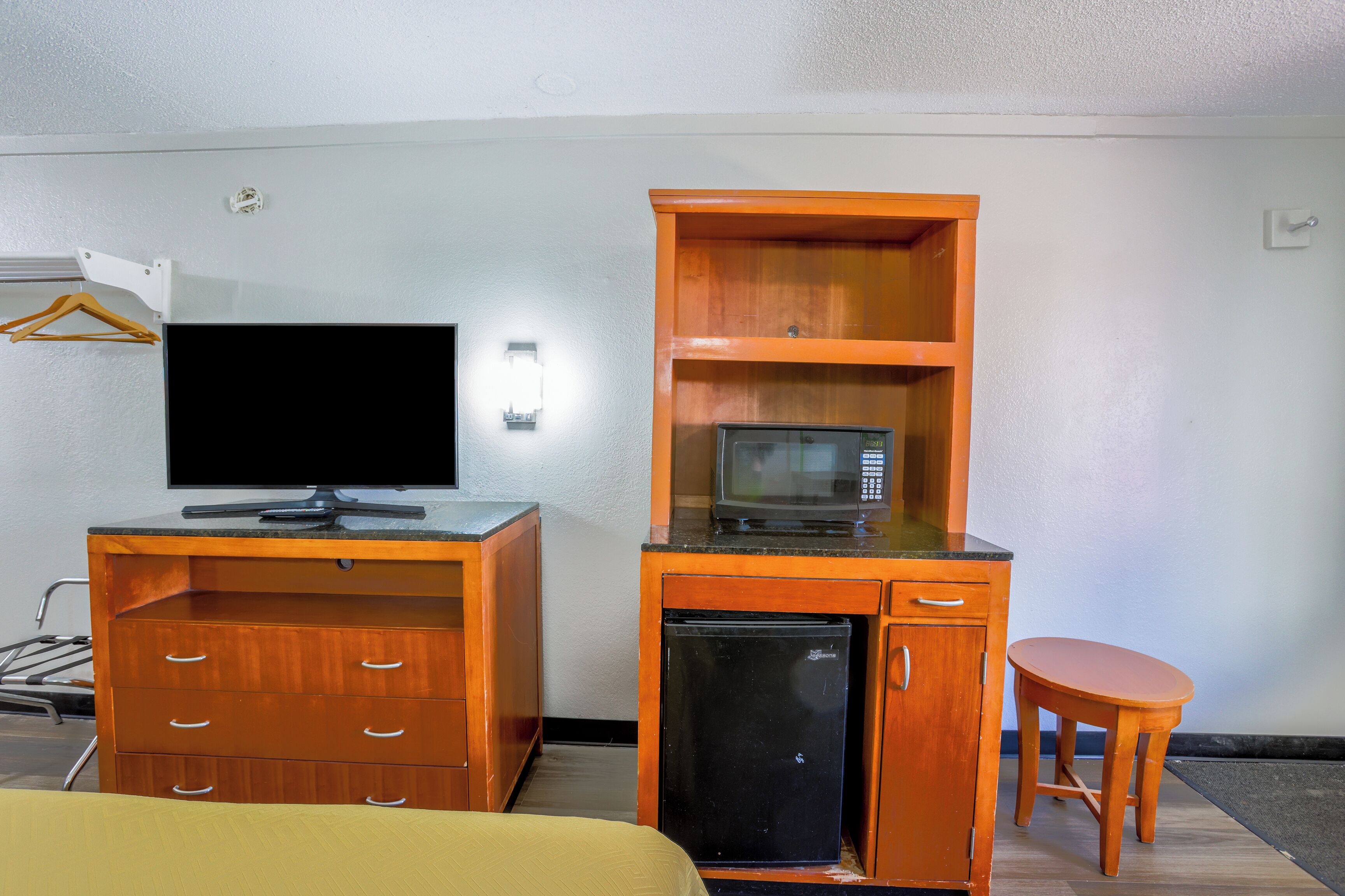 Quarto de hotel compacto com escrivaninha de madeira, televisão, micro-ondas, frigorífico e um pequeno banquinho de madeira.