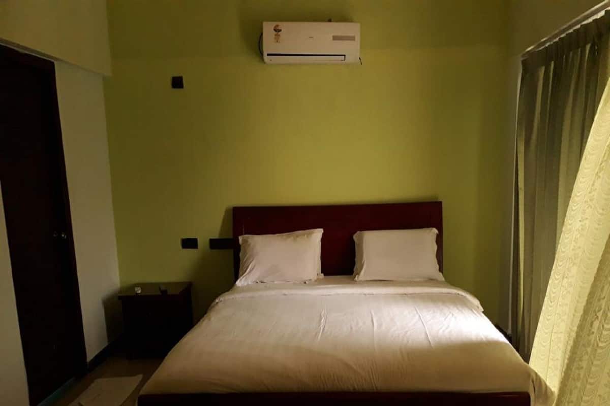A single bed with a wooden headboard in a room with green walls, a small bedside table, and a wall-mounted air conditioner.