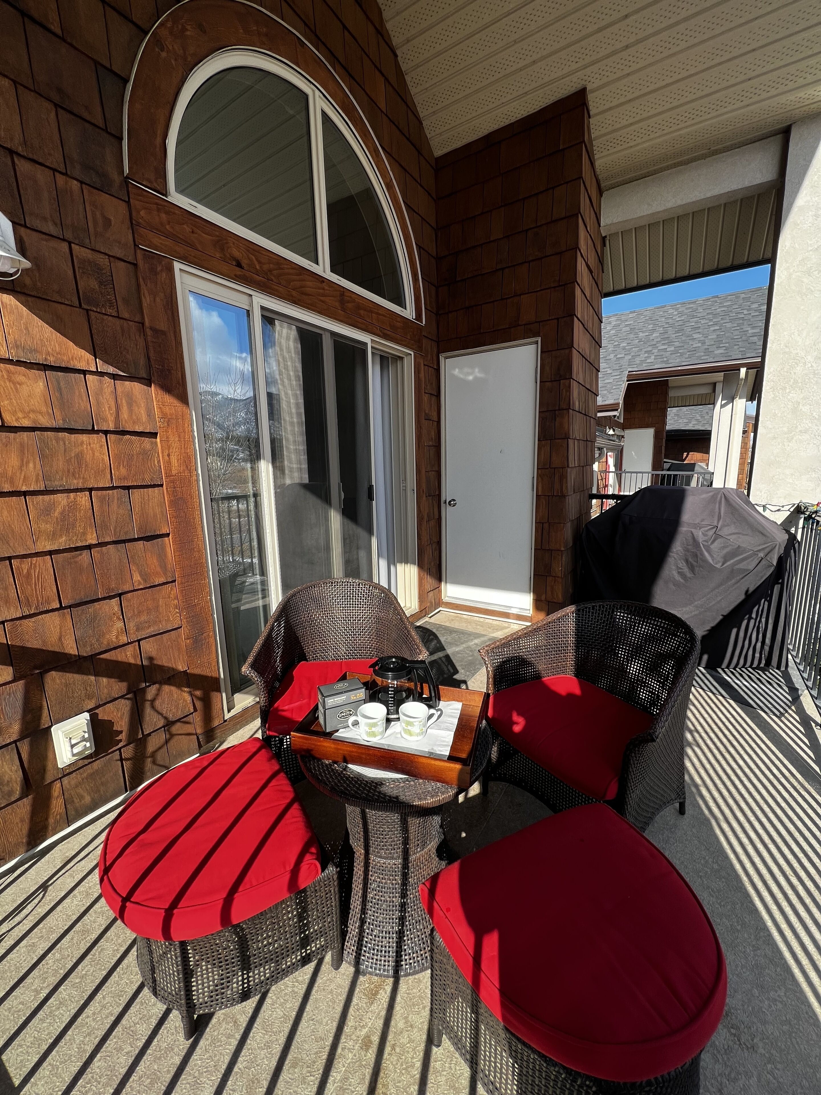 A covered outdoor seating area with wicker furniture and red cushions, a coffee maker, and a tray with cups and a box.