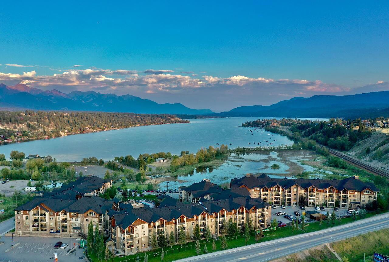 A multi-story hotel with a parking lot, situated near a lake with mountains in the background.