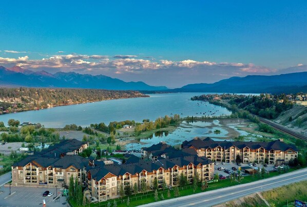 A multi-story hotel with a parking lot, situated near a lake with mountains in the background.