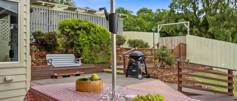 A patio with a checkered tablecloth, a wooden bowl with fruits, a grill, and a bench.
