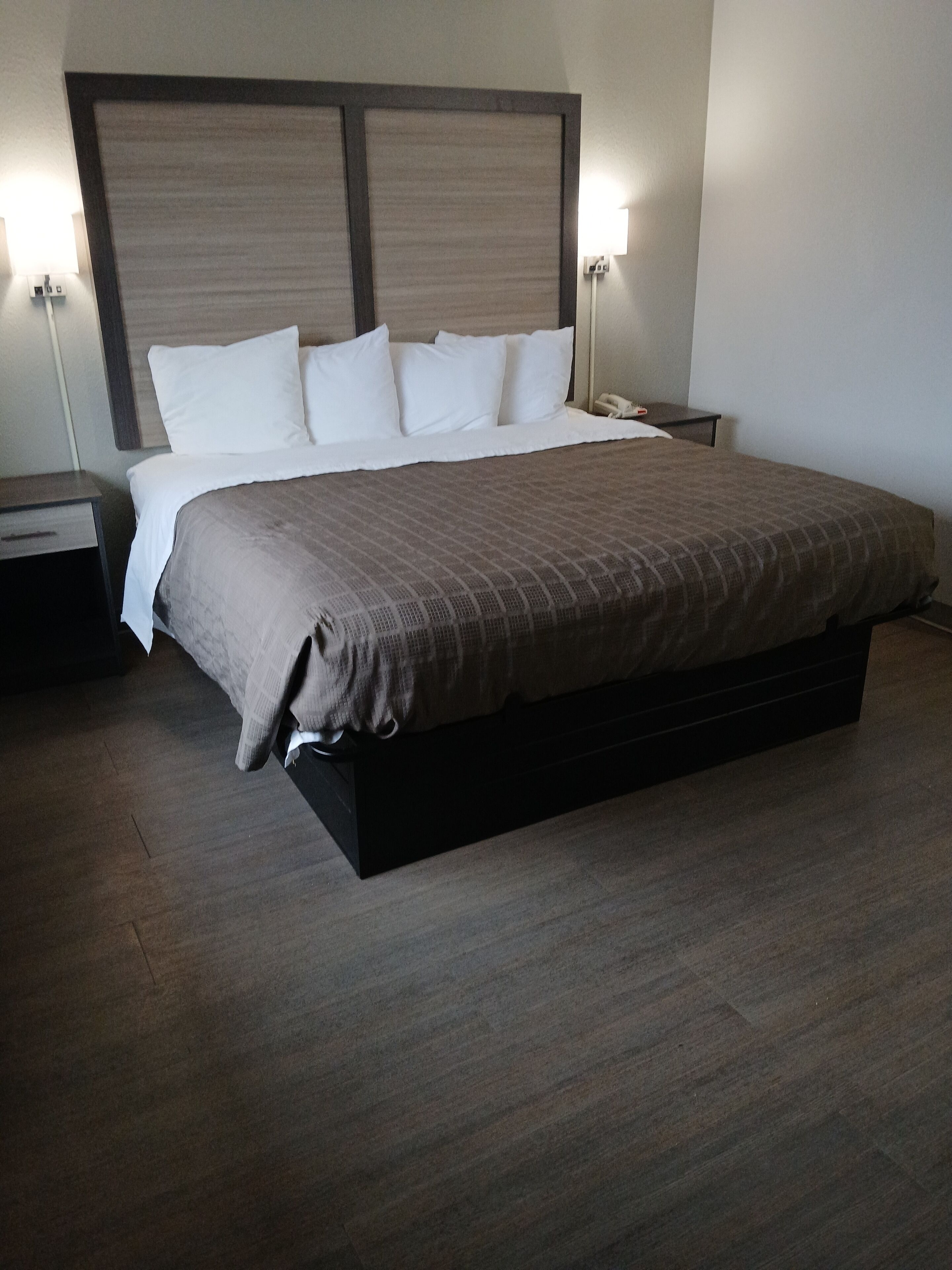 A neatly made bed with a brown bedspread and white pillows, a wooden headboard, two bedside tables with lamps, and a wooden floor.