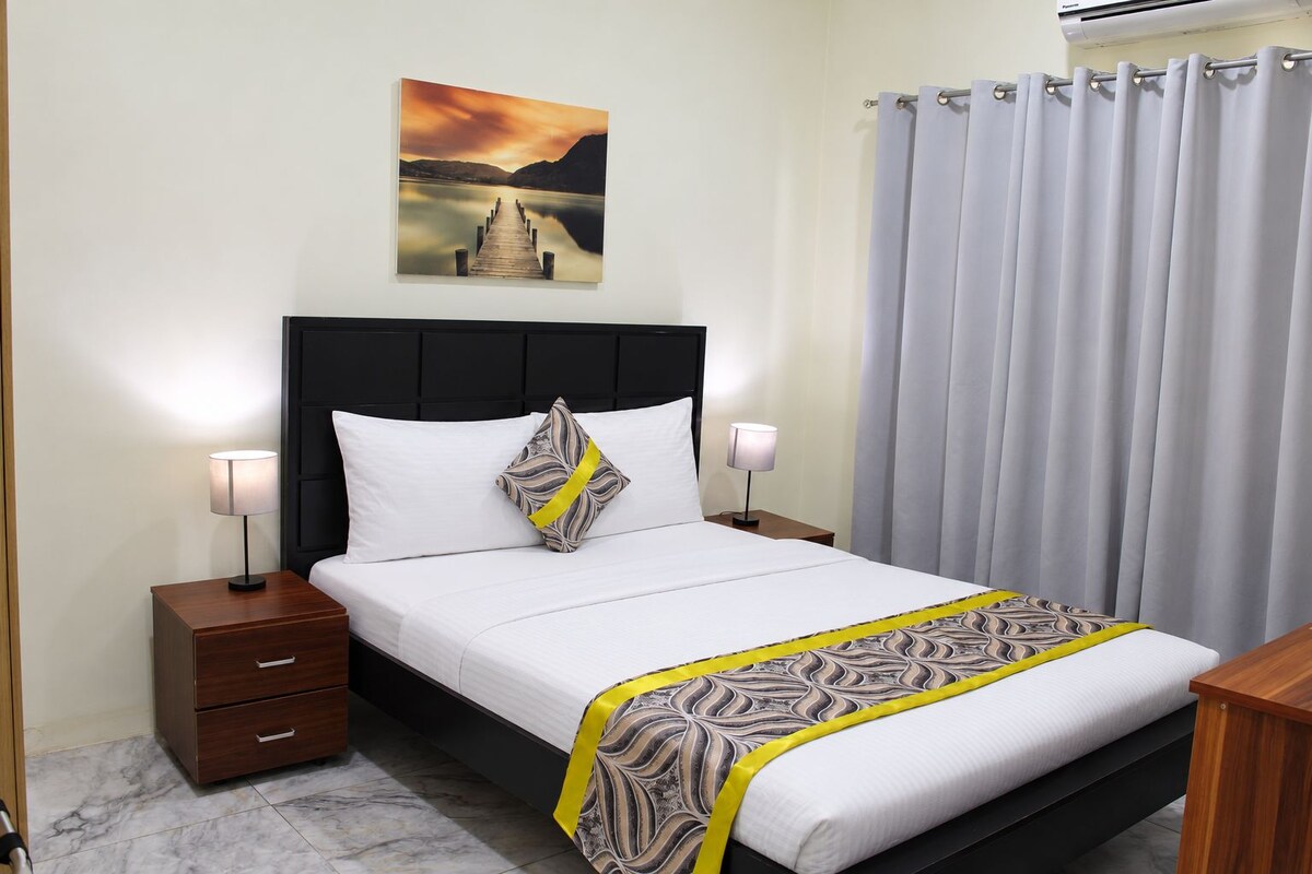 Habitación de hotel con cama, mesitas de noche, lámpara, un cuadro y una cortina.