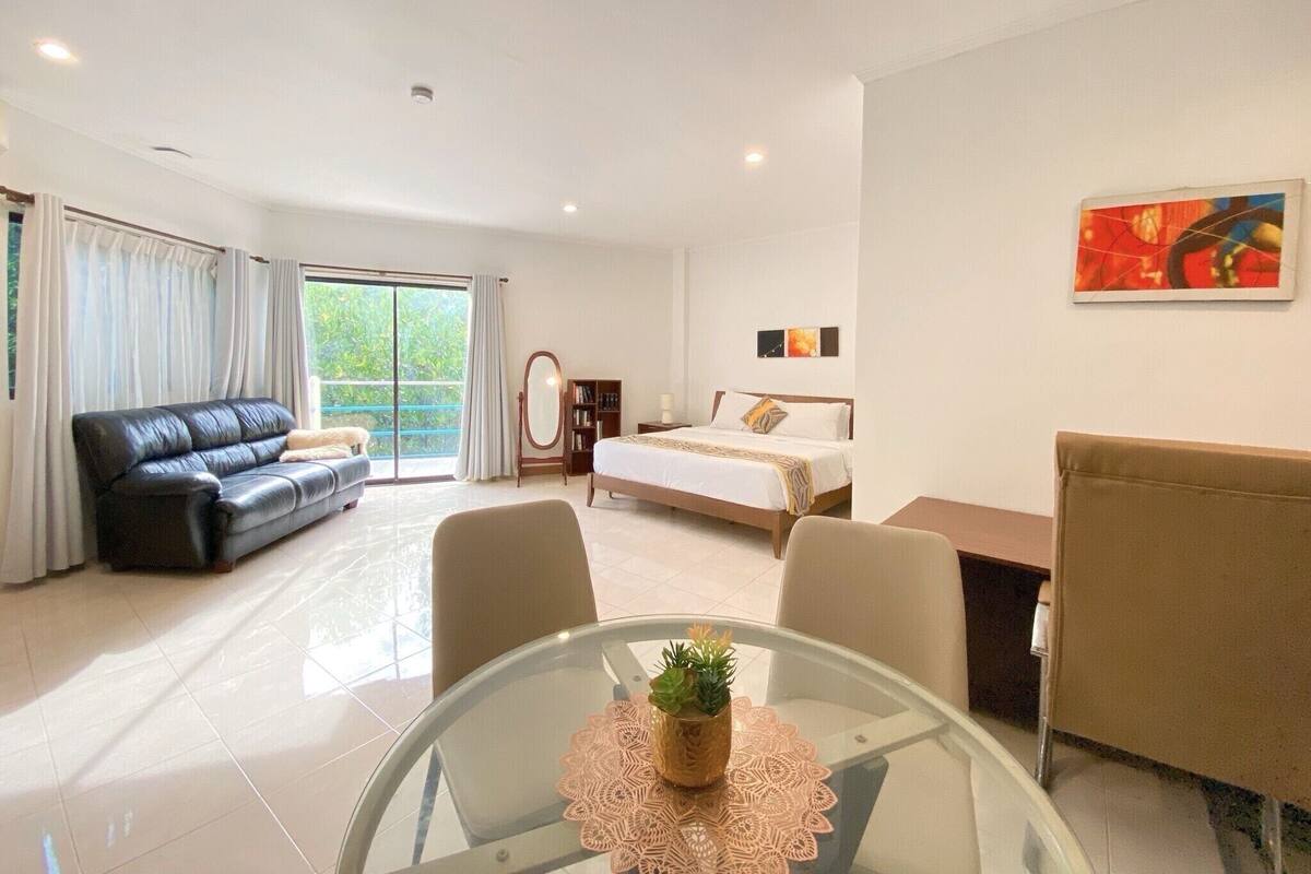 Habitación de hotel moderna con cama, sofá, una mesa de vidrio con una planta y vistas a zonas verdes por la ventana.