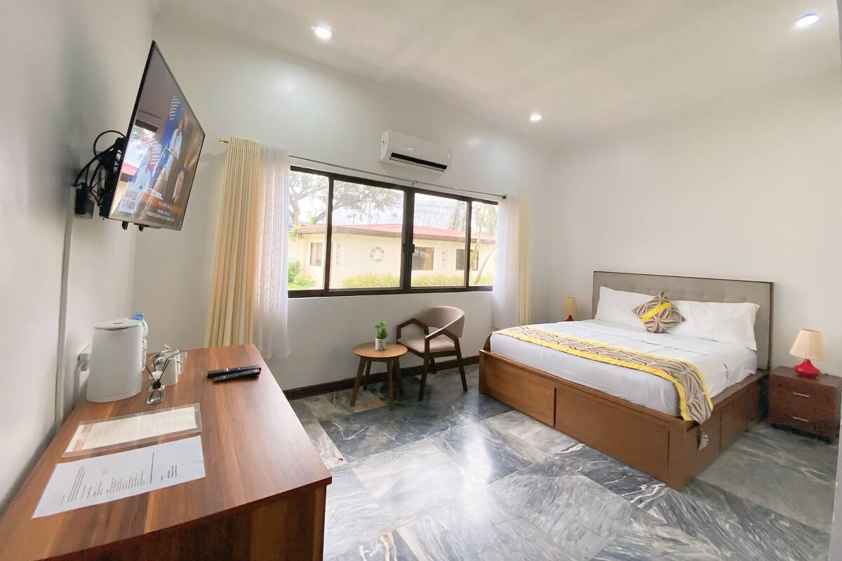 Habitación de hotel con cama, escritorio con papeles, silla, televisor montado en la pared y ventana con vista al exterior.