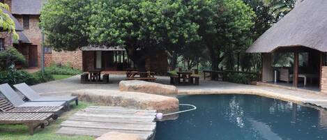 A swimming pool with wooden deck and lounge chairs, surrounded by trees and a brick building.