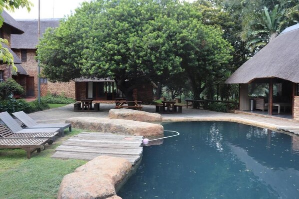 Une piscine avec une terrasse en bois et des chaises longues, entourée d’arbres et d’un bâtiment en briques.