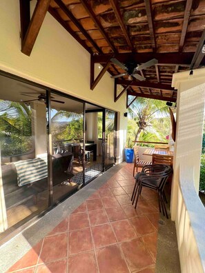 A covered patio with a dining area, large glass doors leading to a view of greenery, and a ceiling fan.