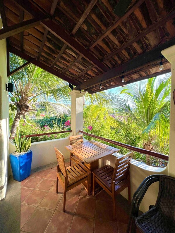 An outdoor patio with a wooden table and chairs, surrounded by lush greenery and a thatched roof.