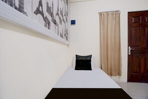 Un lit simple avec un oreiller noir dans une pièce dont le mur est blanc et qui possède une porte.