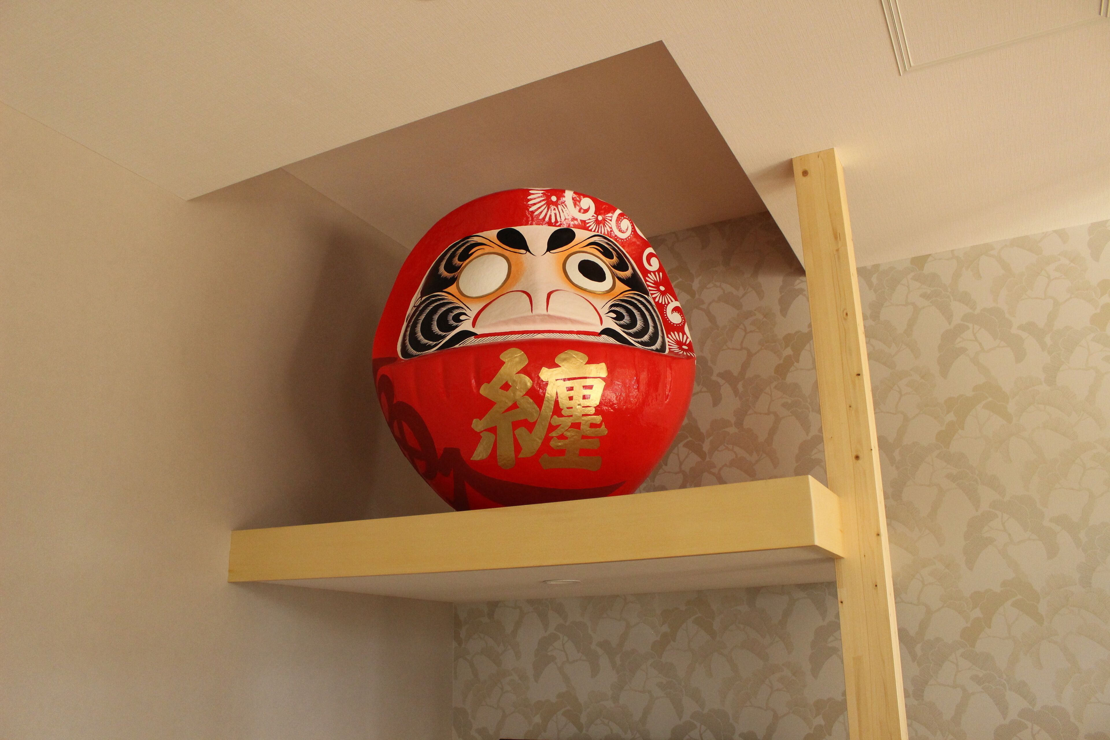 A red decorative object with gold writing on a shelf against a patterned wall.