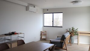 A modern living room with a wooden dining table, a sofa, a small bookshelf, and a large window providing natural light. - Matoi Hostel & Bar (Takasaki)