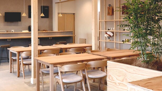A modern interior with wooden tables and chairs, a kitchen area, and a bookshelf. - Matoi Hostel & Bar (Takasaki)