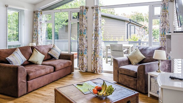 A living room with two brown sofas, a wooden coffee table with fruit, and large windows with curtains.