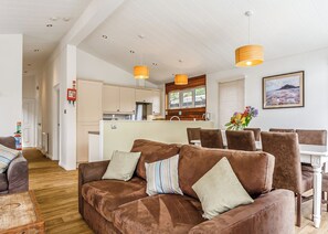 Una sala de estar moderna con un sofá marrón, un comedor con sillas marrones, una cocina con armarios de madera y lámparas colgantes.