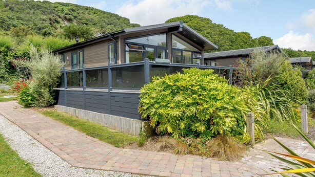 A modern house with large windows, a sloped roof, and a garden with various plants. - Gwithian Premier Lodge (Mullion Cove, Helston)