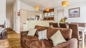 A modern living room with a brown sofa, a dining area with brown chairs, a kitchen with wooden cabinets, and pendant lights. - Gwithian Premier Lodge (Mullion Cove, Helston)