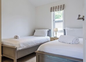 Un dormitorio con dos camas individuales, una ventana con persianas, una mesita de noche con lámpara y una puerta con manija.