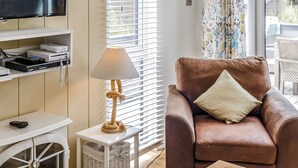 A living room with a brown leather sofa, a white side table, a lamp, a television on a shelf, and a wicker basket. - Harbour Premier Lodge (Mullion Cove, Helston)