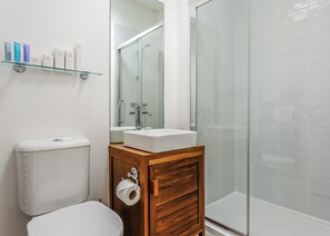 A bathroom with a toilet, a sink with a mirror, a glass shower enclosure, and toiletries on a shelf.