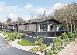 A modern mobile home with a balcony, surrounded by landscaped garden beds and a paved driveway. - Constantine Premier Lodge (Mullion Cove, Helston)