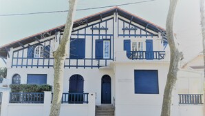 Un edificio de dos pisos con fachada blanca y detalles azules, que cuenta con un balcón en el piso superior y una puerta principal con unos escalones que la anteceden.