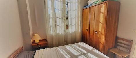 A bedroom with a bed, a wooden wardrobe, a chair, a nightstand with a lamp, and a window with sheer curtains.