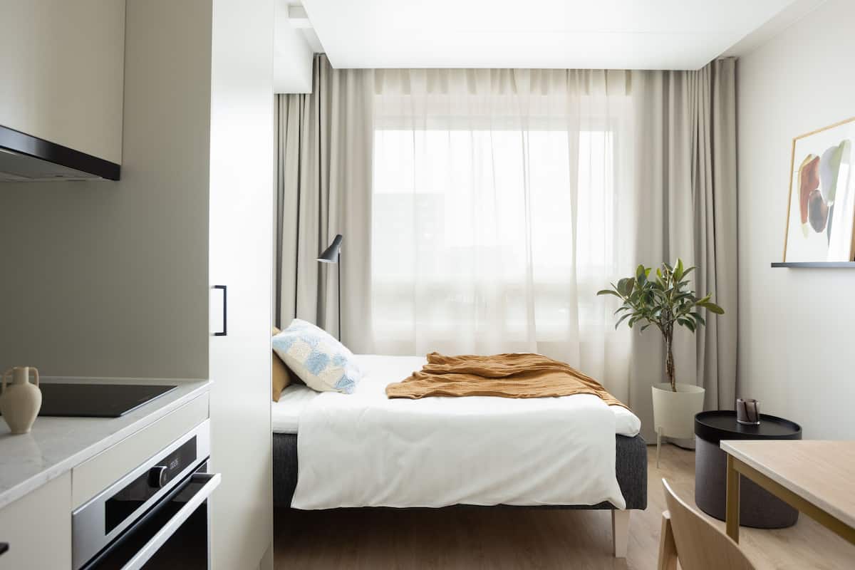 A modern hotel room with a bed, a dining table, a potted plant, and a kitchenette.