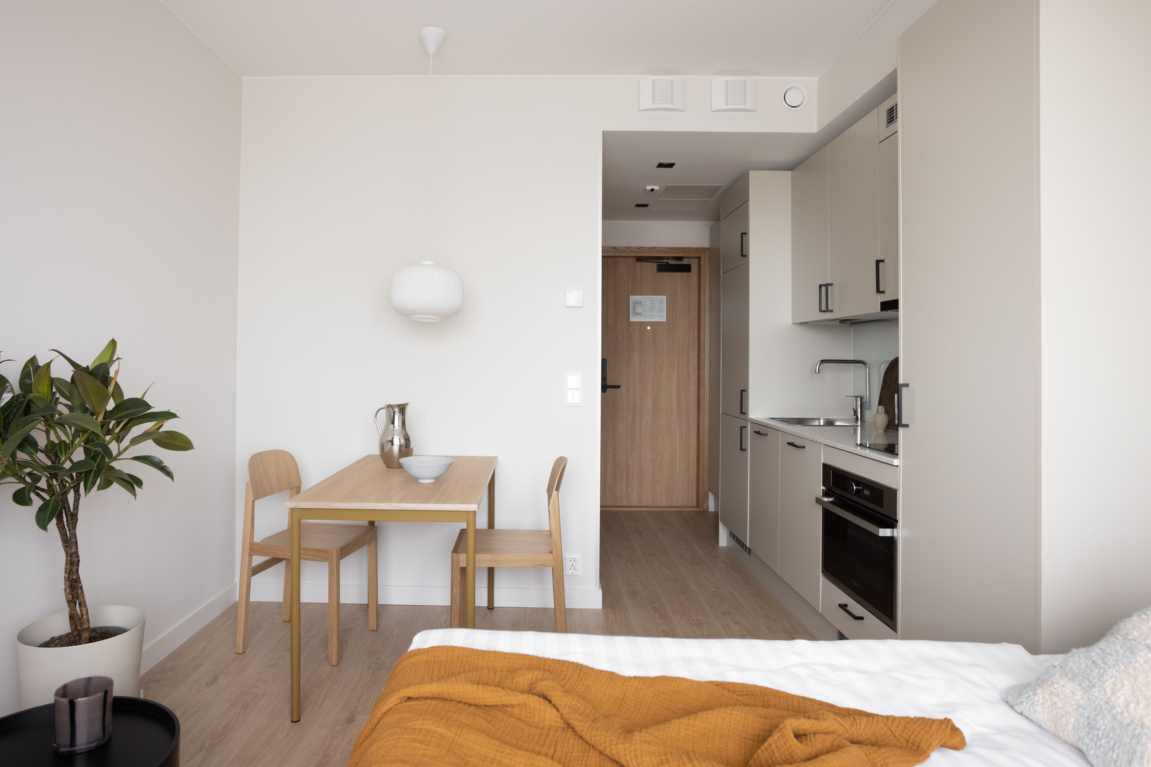 Een moderne, compacte hotelkamer met een bed, een klein eettafeltje, stoelen, een potplant en een kitchenette.