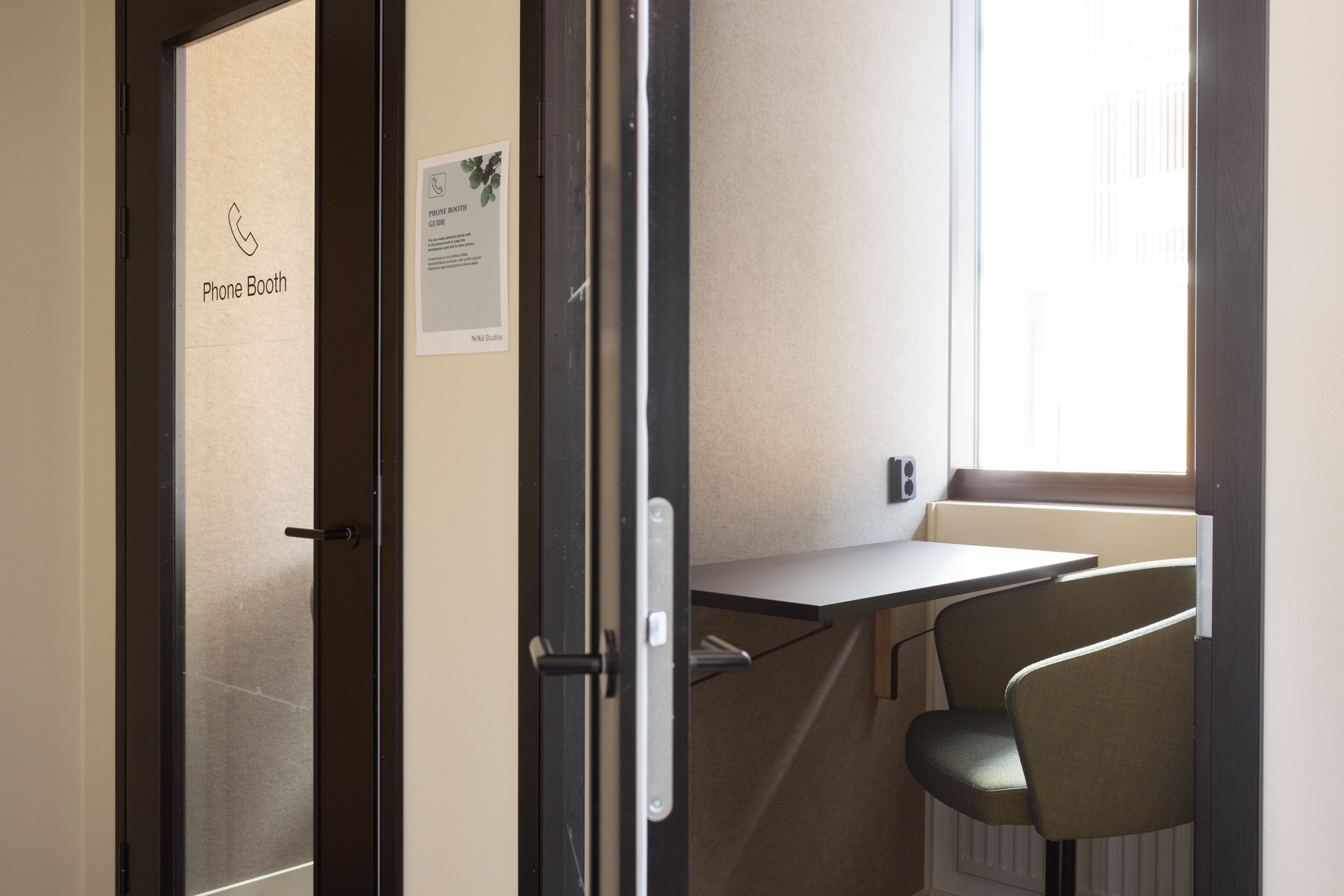 A phone booth with a sign, a small table, and a single chair in a room with a window.