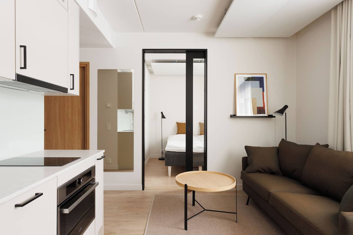A modern hotel room with a kitchenette, a sofa, a small round table, and a bed in the background.