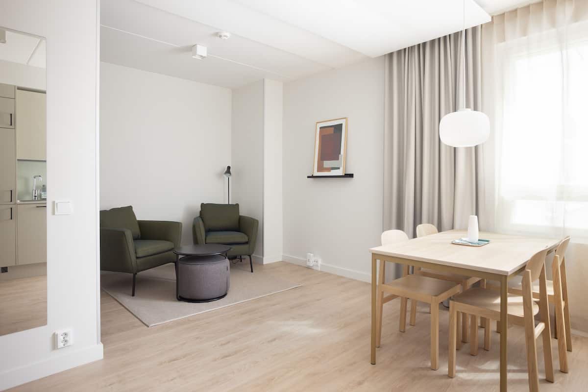 A modern living room with a dining area, featuring a light wooden floor, green armchairs, a round coffee table, and a white table with wooden chairs.
