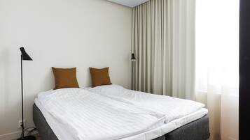 A neatly made bed with white linens and two brown pillows, a black floor lamp, and a window with sheer curtains.