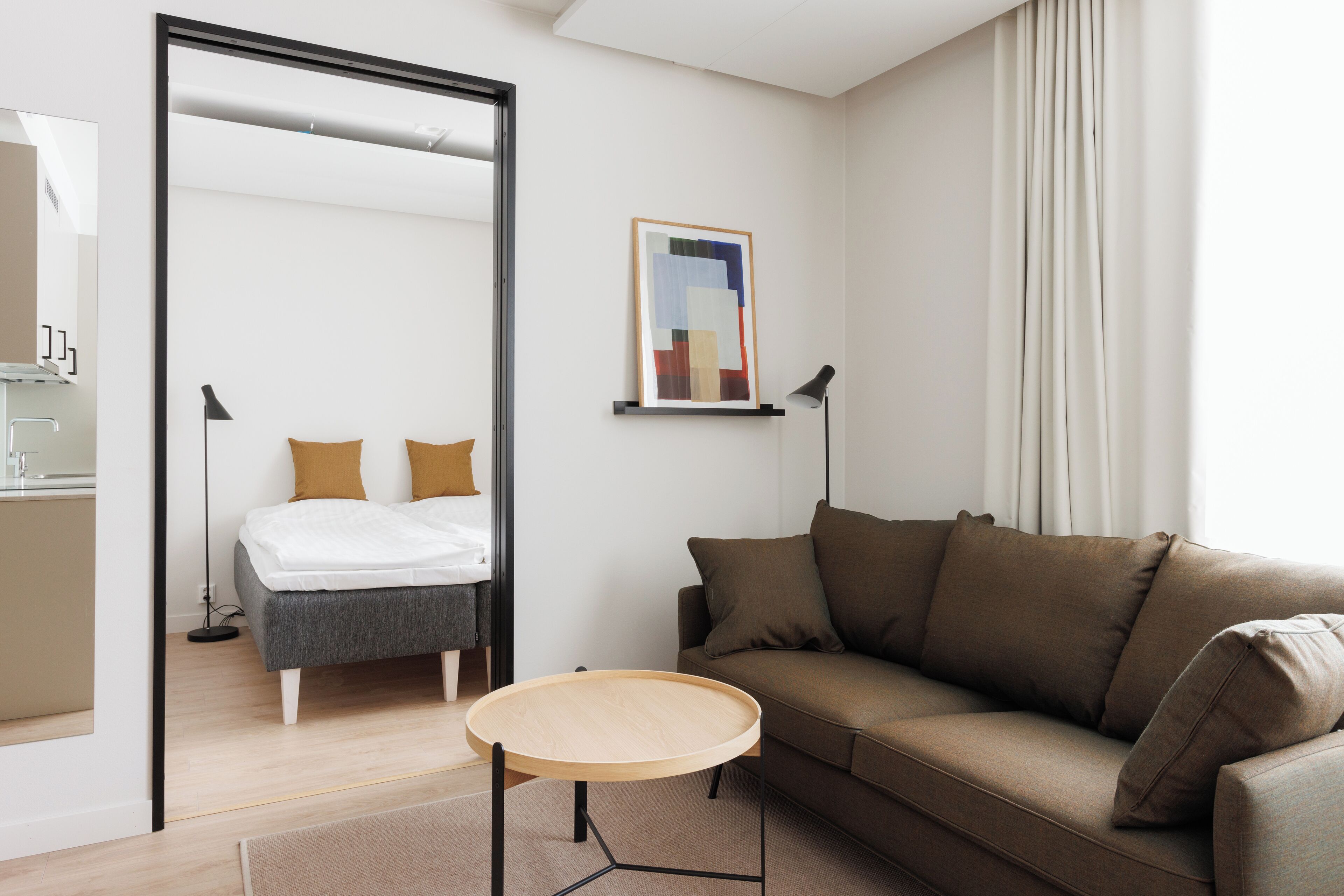 Moderni hotellihuone, jossa on sänky, ruskea sohva, pyöreä puinen sivupöytä ja taulu seinällä.