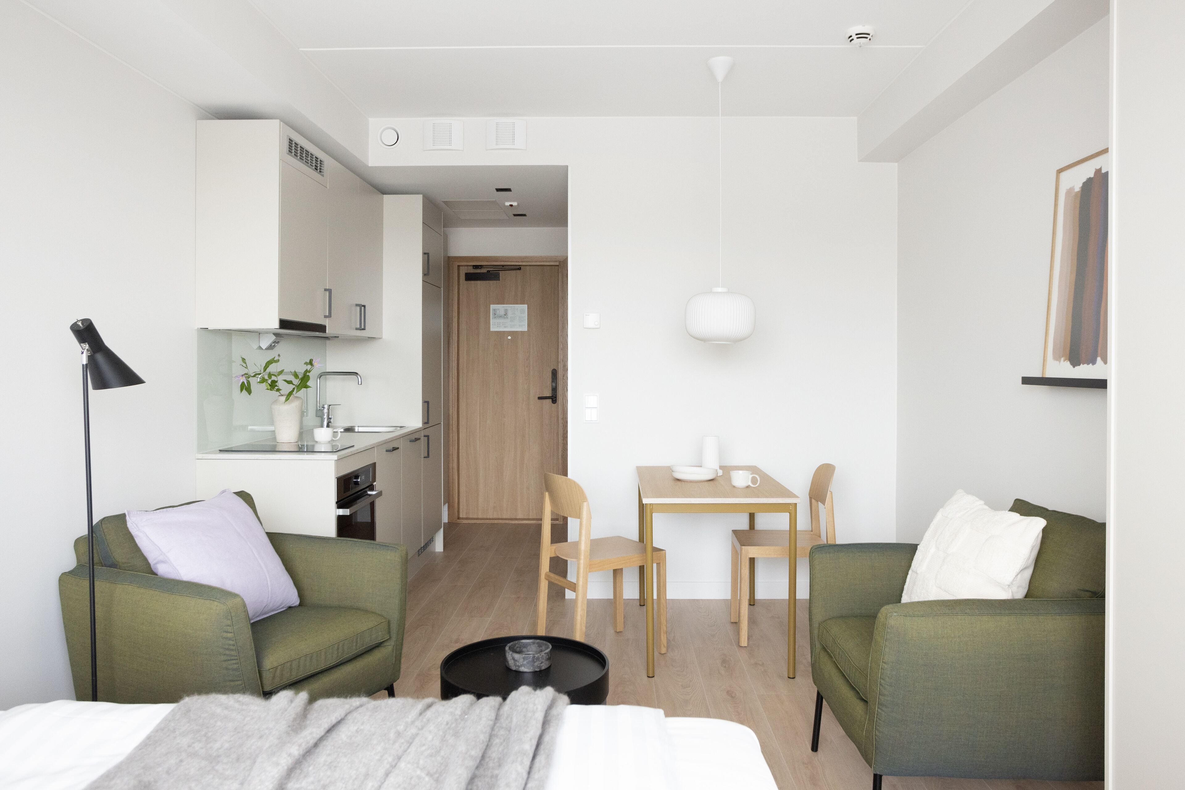 A modern hotel room with a green sofa, a small dining table, a coffee table, and a kitchenette in the background.