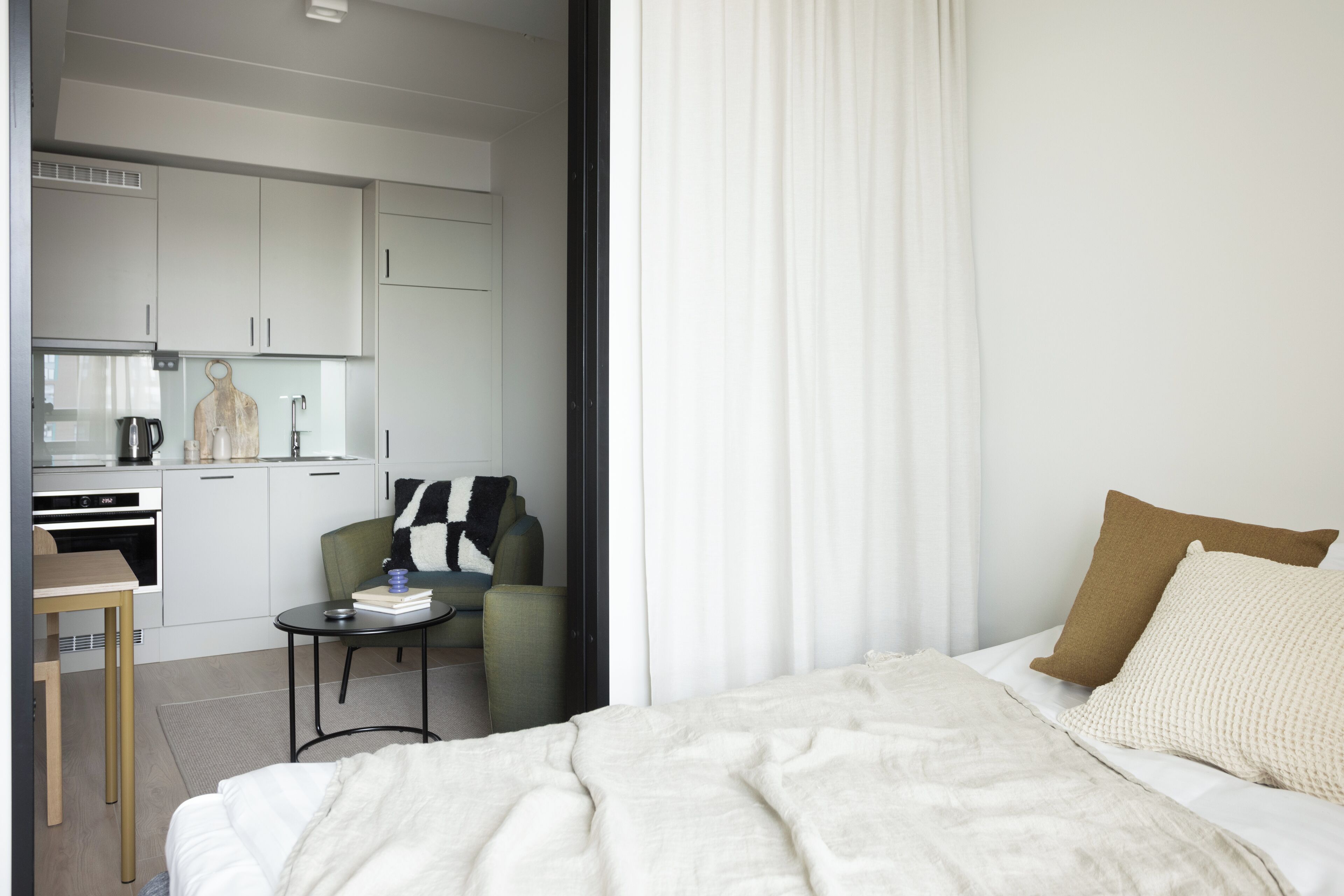 A modern hotel room with a bed, a small table with a book, a chair, and a kitchenette area.