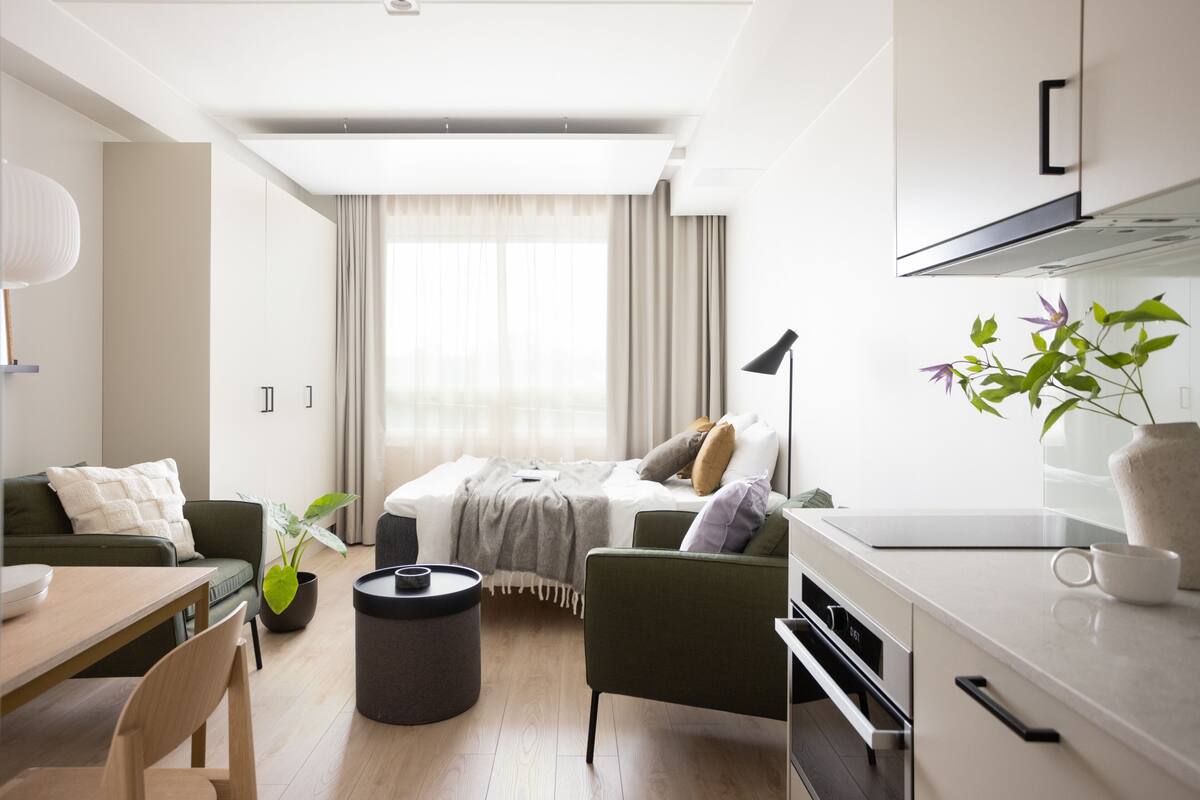 A modern hotel room with a sofa, a dining table, a small kitchen area, and a large window with curtains.