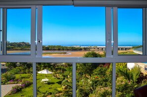 Vista a través de una ventana de un jardín exuberante con un patio, una playa de arena y un puente a lo lejos.