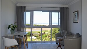 A modern living room with a large window, a dining table, and two sofas. - Serenity Ocean Lodge (Wilderness)
