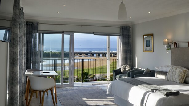 A hotel room with a large bed, a desk, and a chair. There is a view of the beach and the ocean through the glass doors. - Serenity Ocean Lodge (Wilderness)