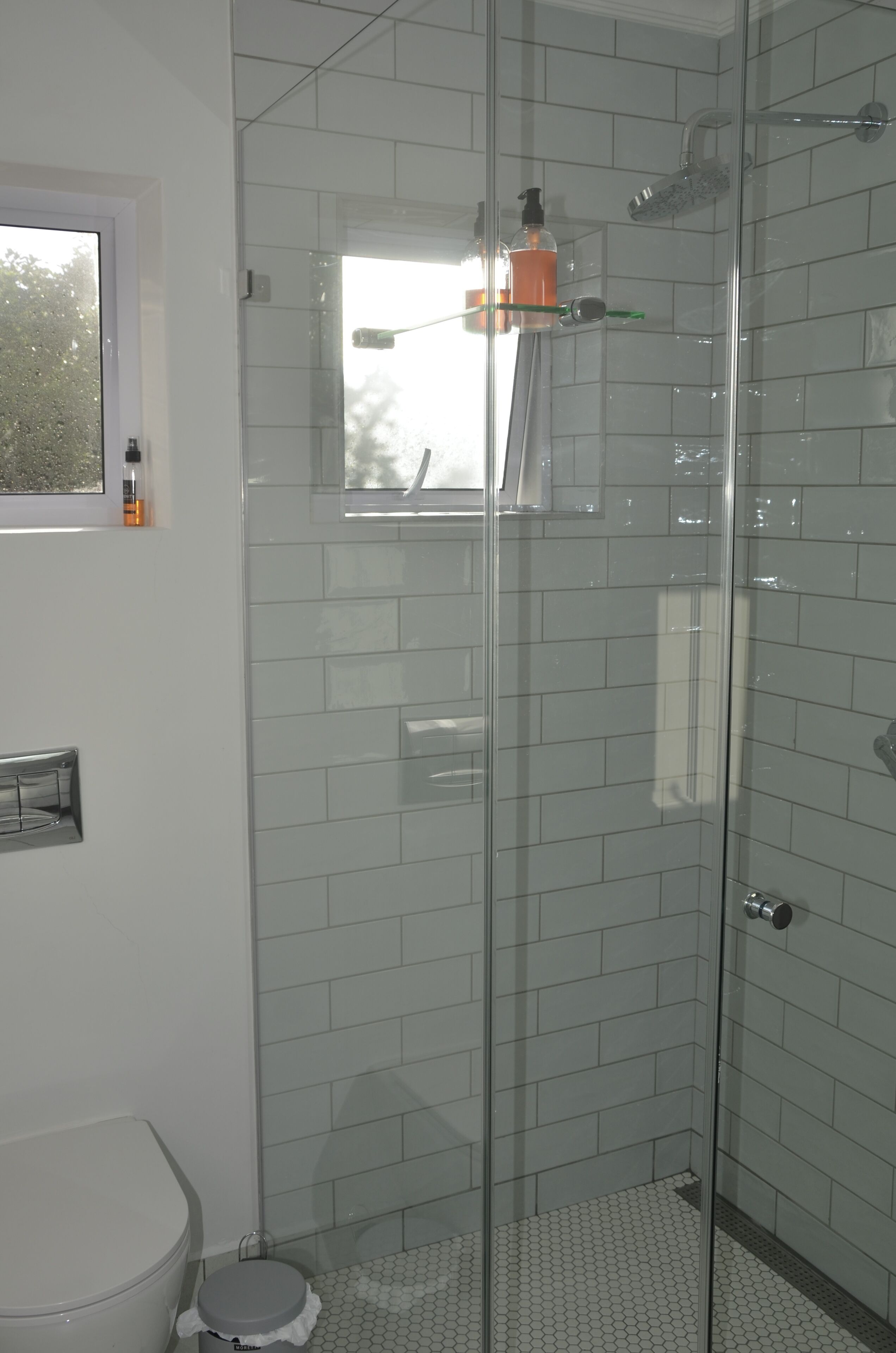 A modern bathroom with a glass shower enclosure, white tiled walls, and a small window.