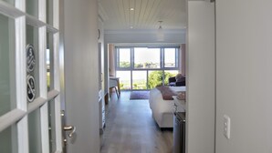 A modern, well-lit hallway leading to a bedroom with a large window, a bedside table, and a chair. - Serenity Ocean Lodge (Wilderness)