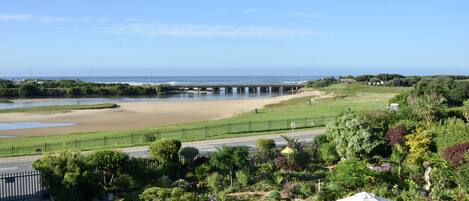 A coastal view with a beach, a bridge, and a garden with a table and chair.