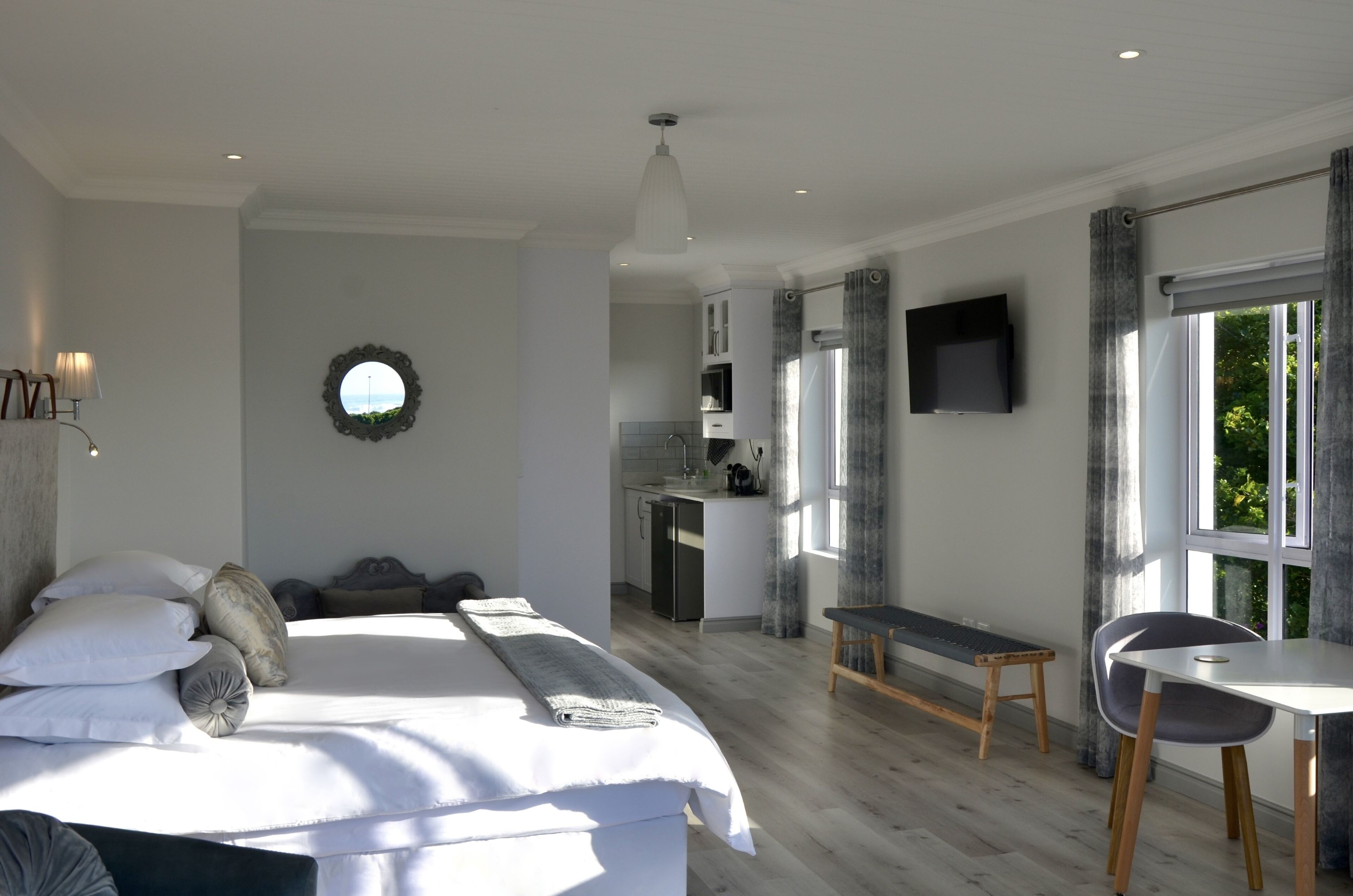 Une chambre à coucher avec un lit, une table de chevet et un petit bureau avec une chaise. Il y a une fenêtre avec des rideaux, un téléviseur fixé au mur et, au fond, un coin cuisine.
