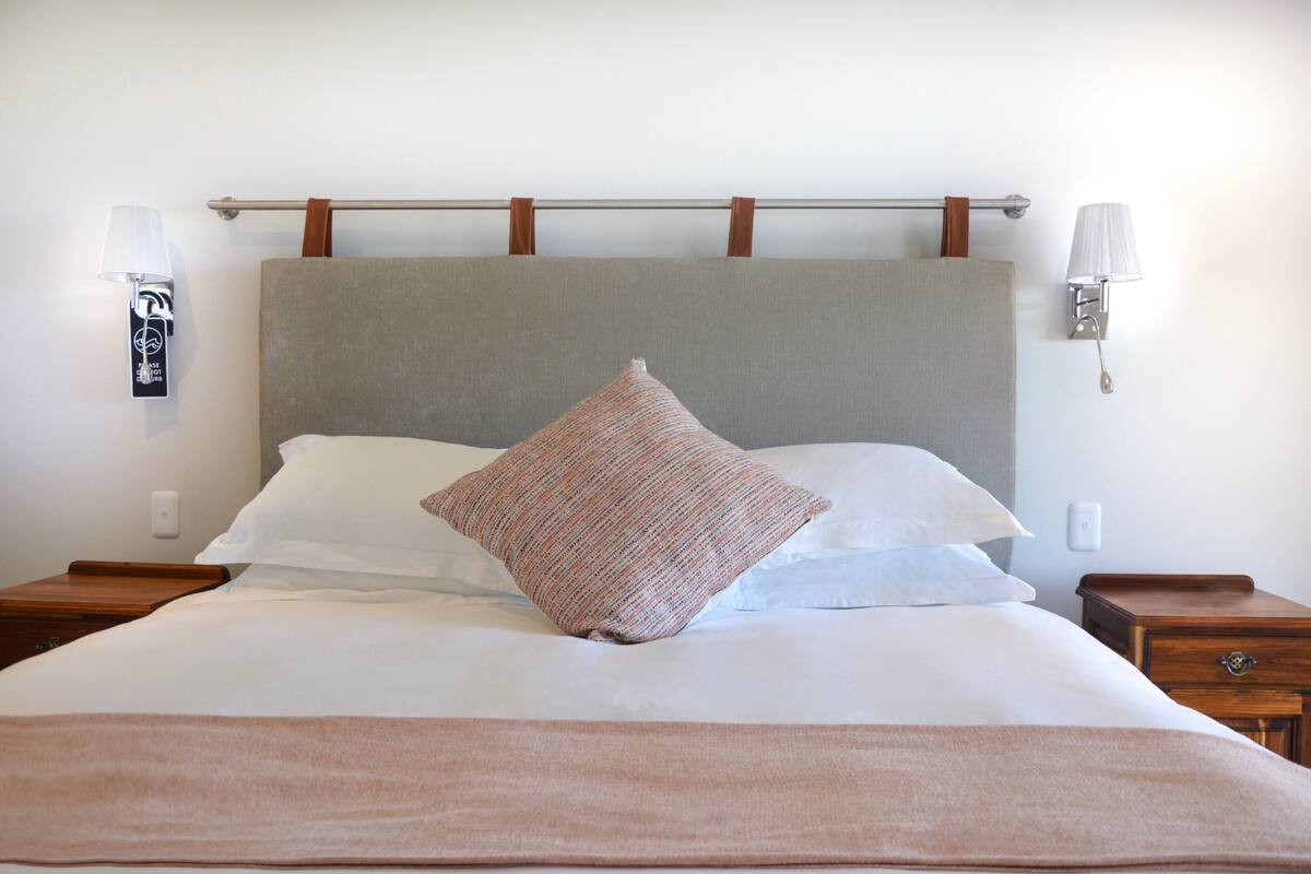 A neatly made bed with a grey headboard, white pillows, and a brown accent pillow. There are two bedside tables with lamps and a wall-mounted light fixture.